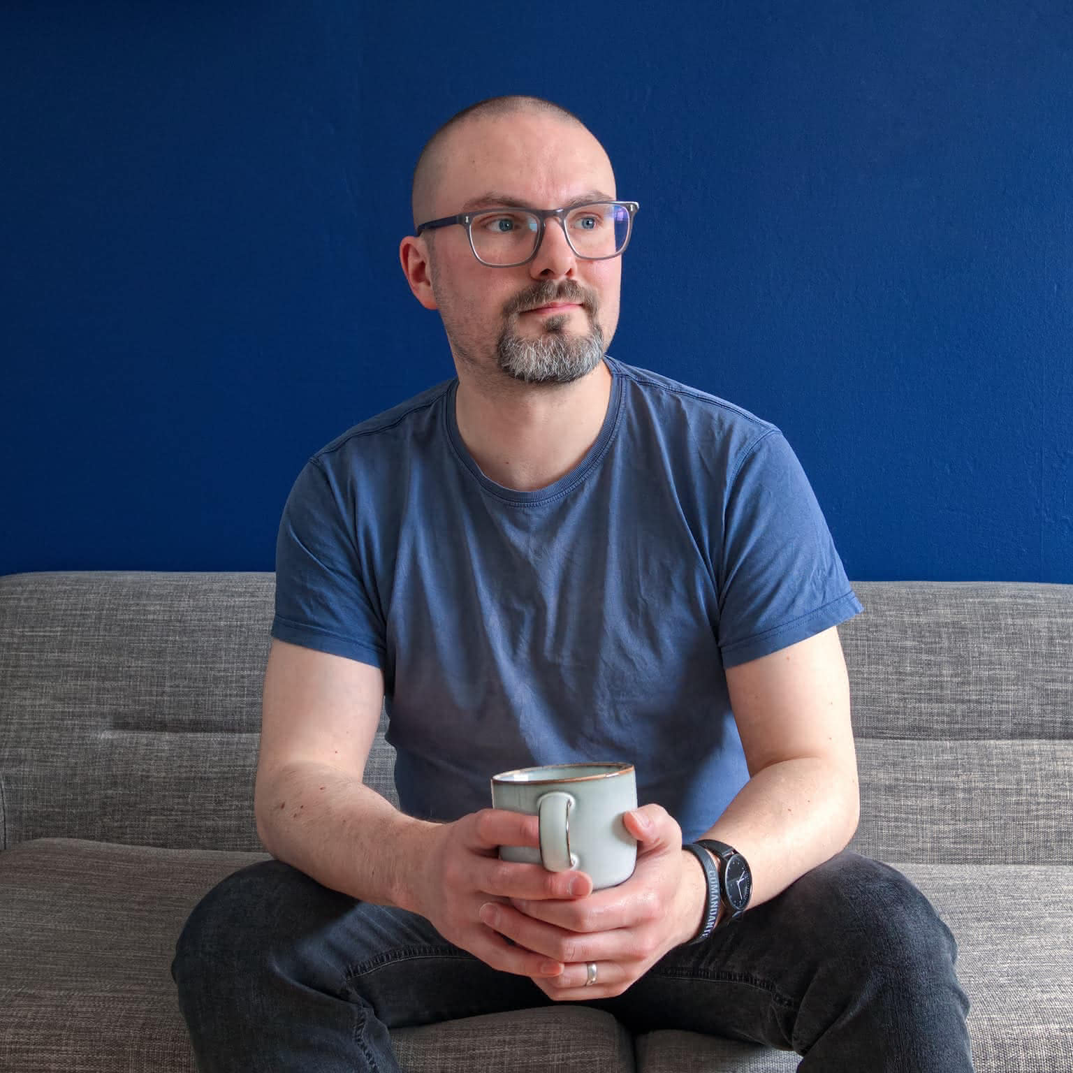 Ryan sat on a grey couch, holding a mug. He wears grey glasses and a blue t-shirt, with a blue wall in the background.